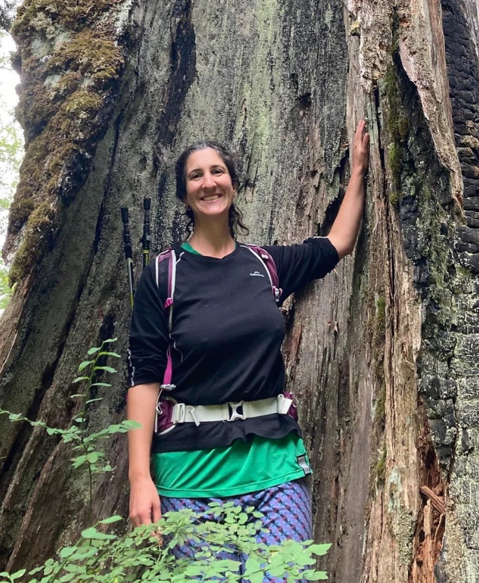 Elana standing beside the trunk of an enormous moss-covered tree, one hand resting on the bark